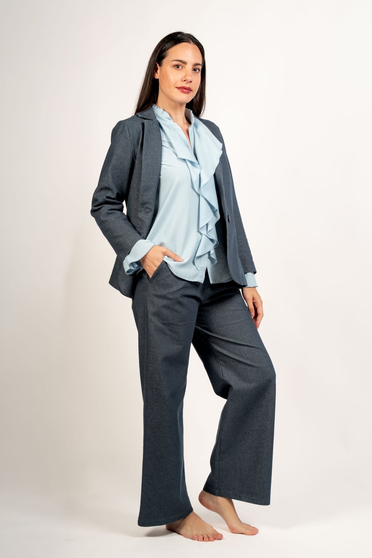 Pantalón de pernera ancha vaquero azul con americana y blusa de volantes, mano en el bolsillo