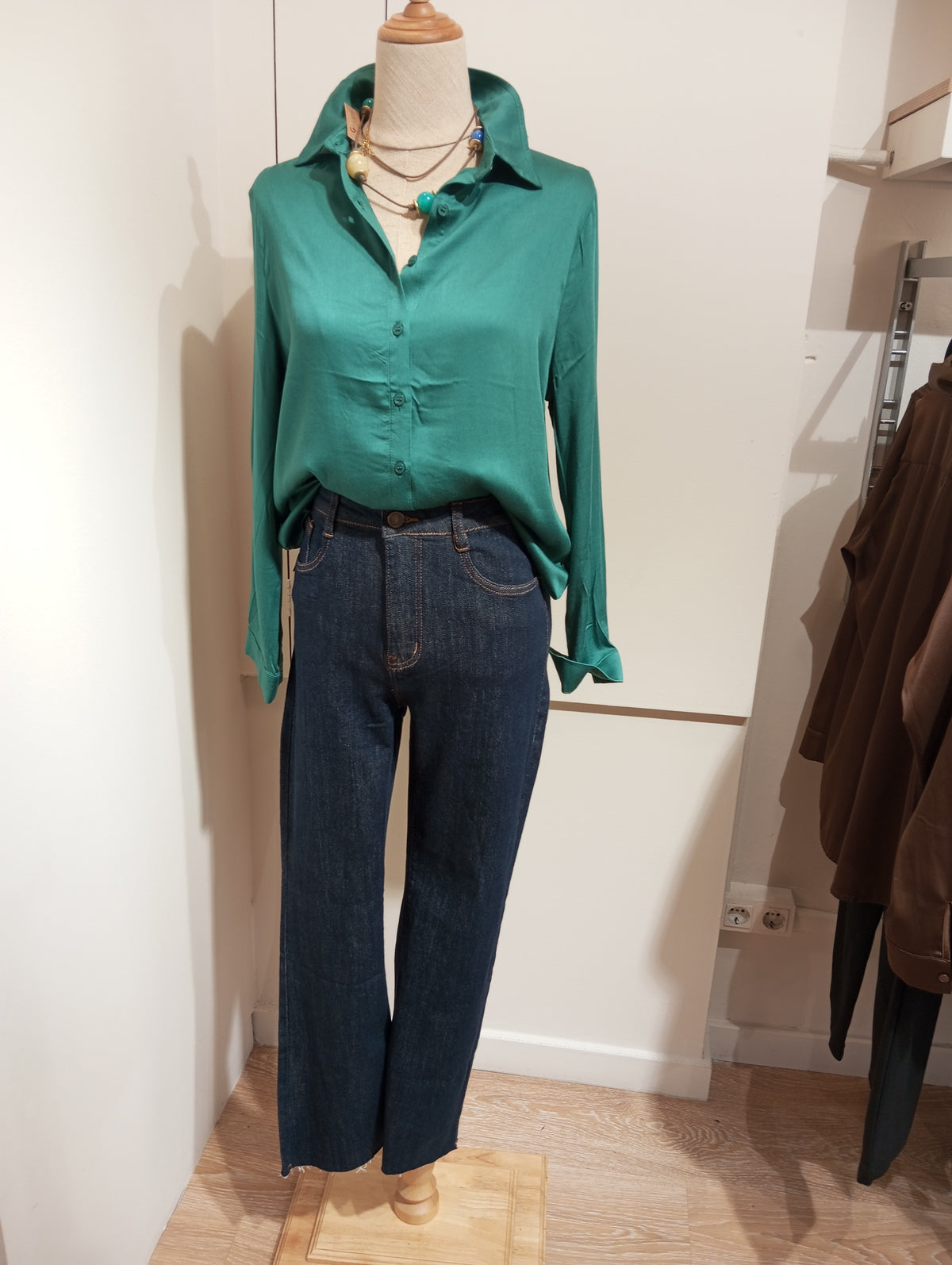 Camisa satinada verde con tejanos oscuros y collares superpuestos, en maniquí