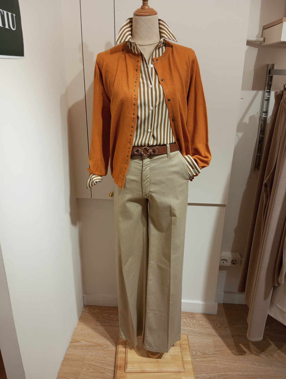 Rebeca mostaza con tachas sobre camisa de rayas, cinturón marrón y pantalón beige, en maniquí